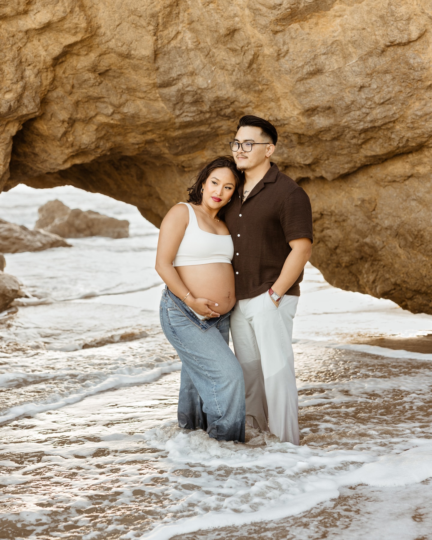 A stylish couple in casual denim posing by the coastal rock cliffs during a Malibu beach maternity photography session.