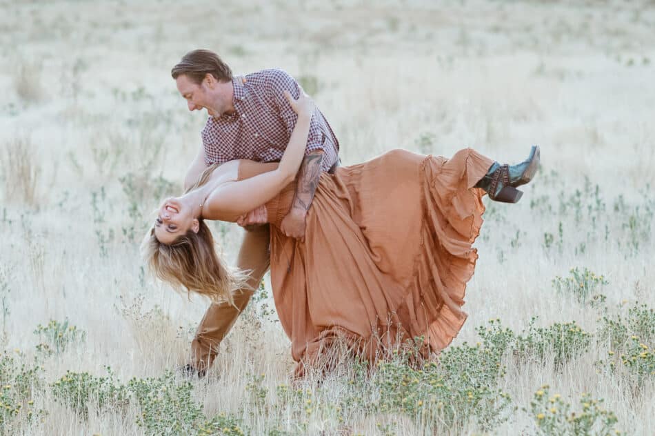 Couple laughing as man dips woman in a field during engagement session with natural light