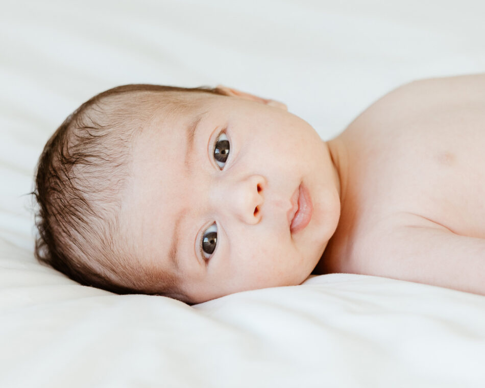 A close-up lifestyle newborn photography portrait highlighting sharp details and soft skin tones in Calabasas, California. An alert newborn baby with fine dark hair lies comfortably on a crisp white blanket, looking directly into the camera.