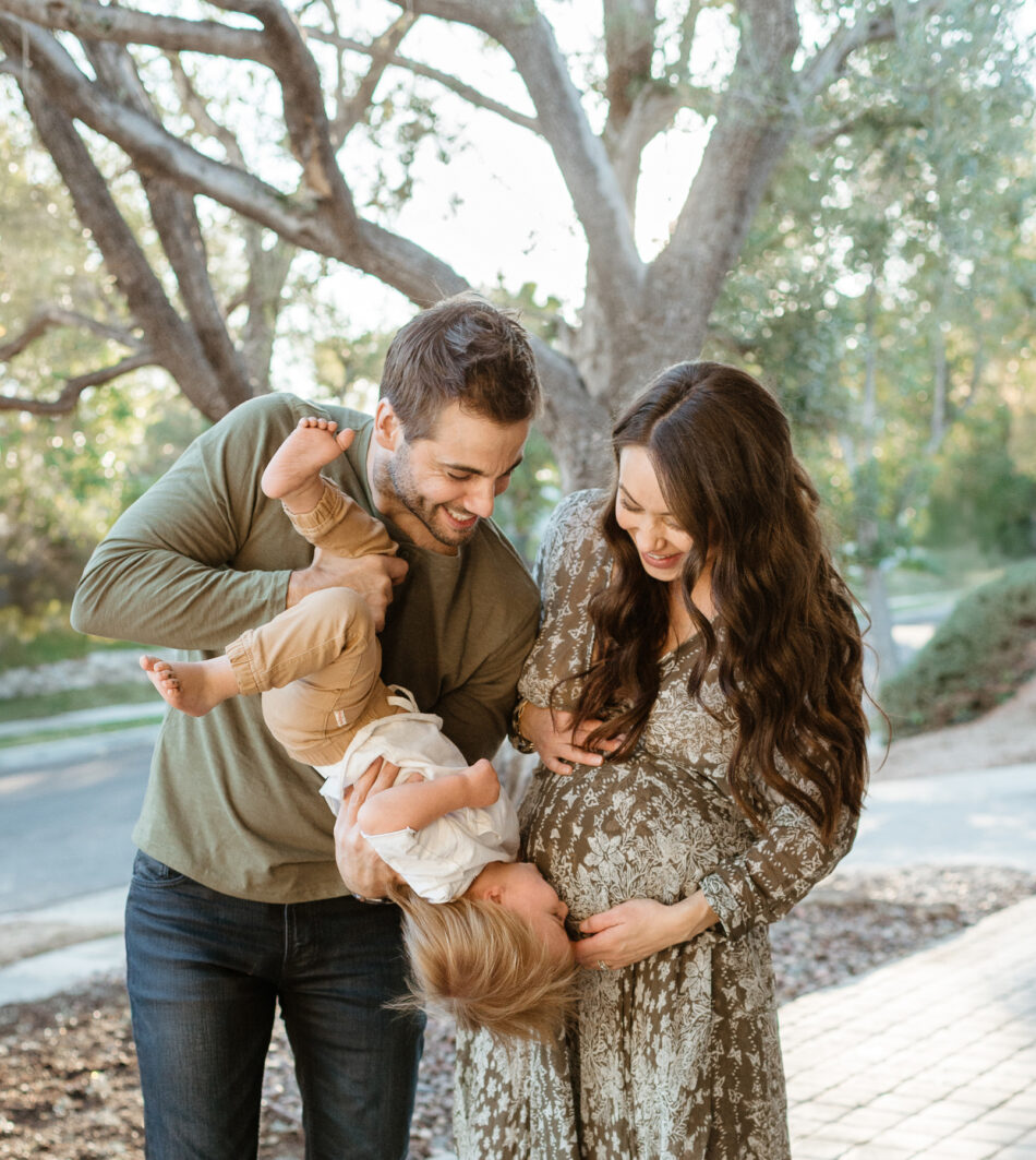 Parents laughing while holding their toddler upside down next to a pregnant mother during a lifestyle maternity photography session at home photographed by Erica Montgomery.