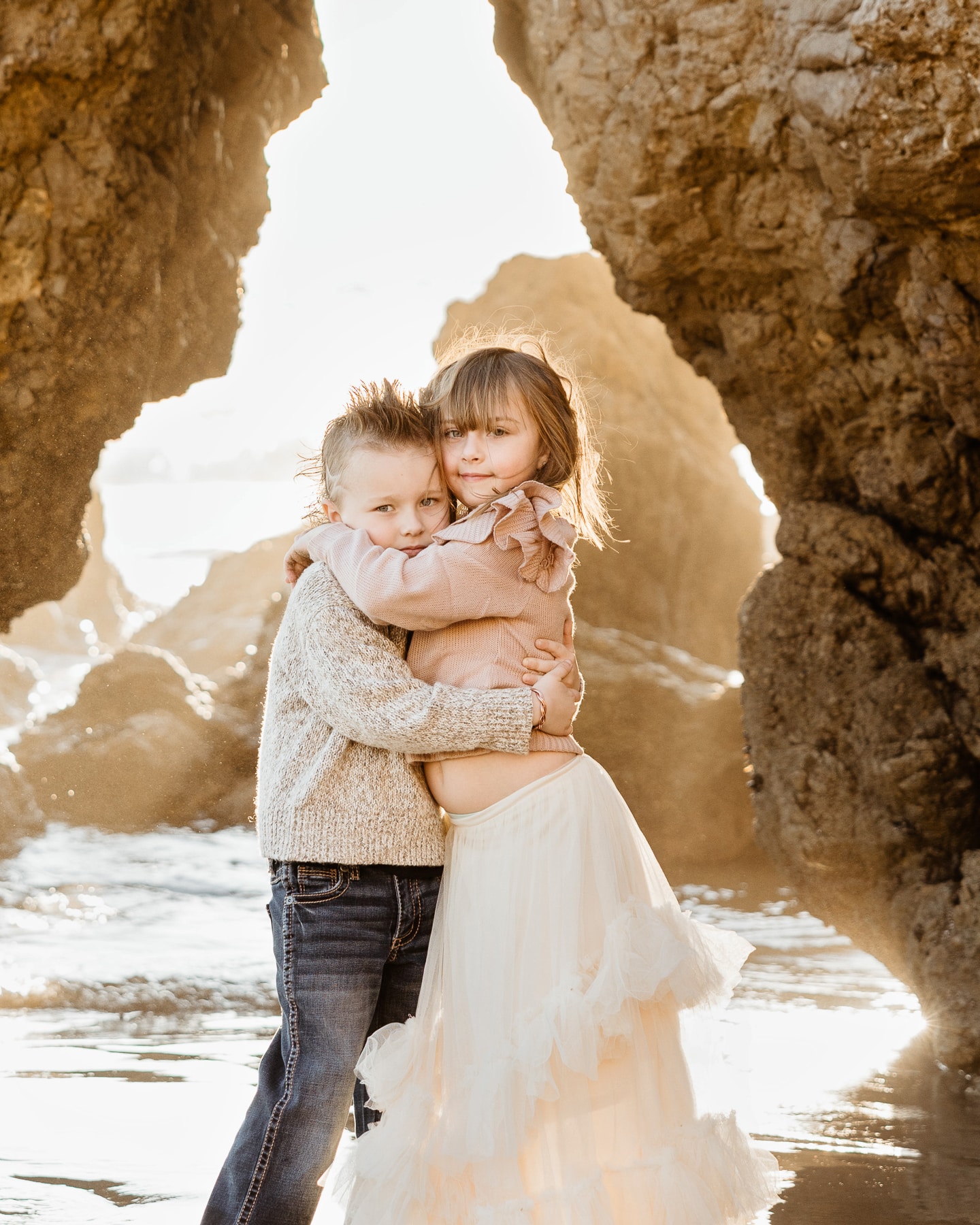 Two young siblings hugging tightly inside a rocky sea cave during a golden hour Malibu beach family photography session by Erica Montgomery Photography.