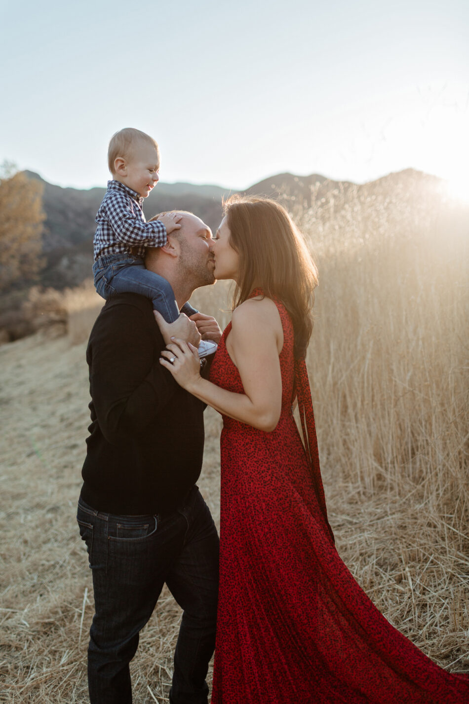 Parents kissing in a sunlit canyon field while their toddler sits on dad's shoulders during a Calabasas family photoshoot by Erica Montgomery Photography.