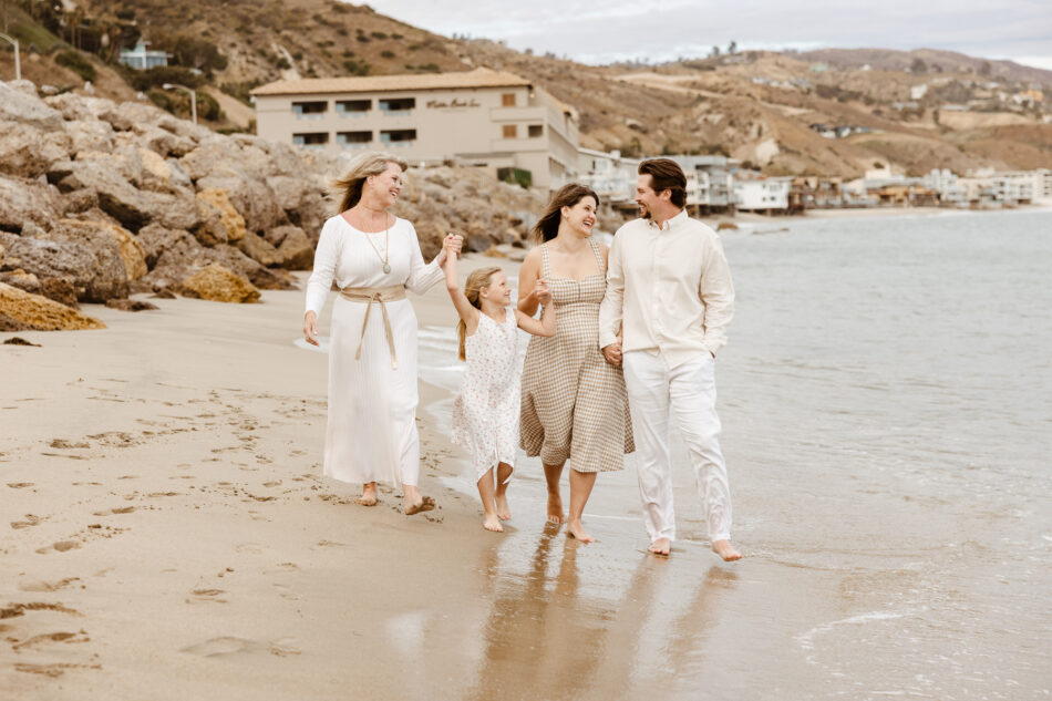 Luminous editorial photo of a family walking along the water's edge near a rocky Malibu coastline backdrop and the Malibu Beach Inn.
