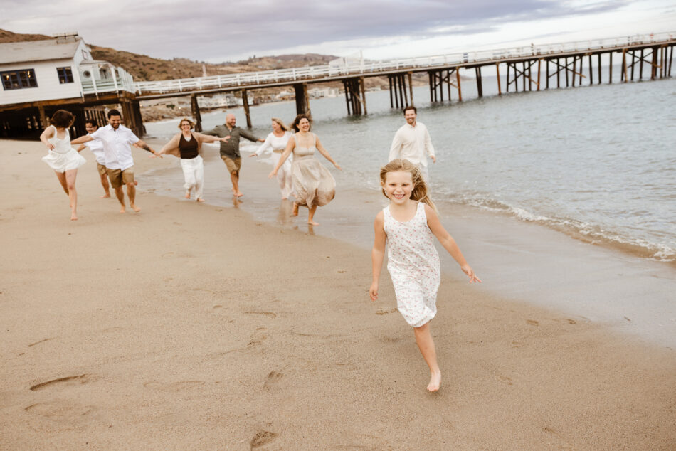 Joyful candid photo of a young girl running down the sand with her extended family following closely behind near the Malibu Pier.
