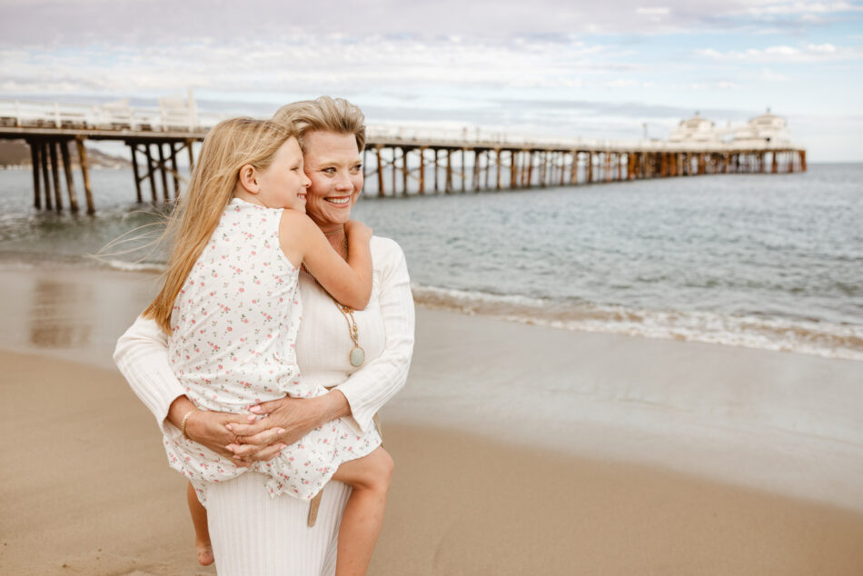 Heartfelt candid photo of a grandmother hugging her young granddaughter tightly on the shoreline with the Malibu Pier behind them.
