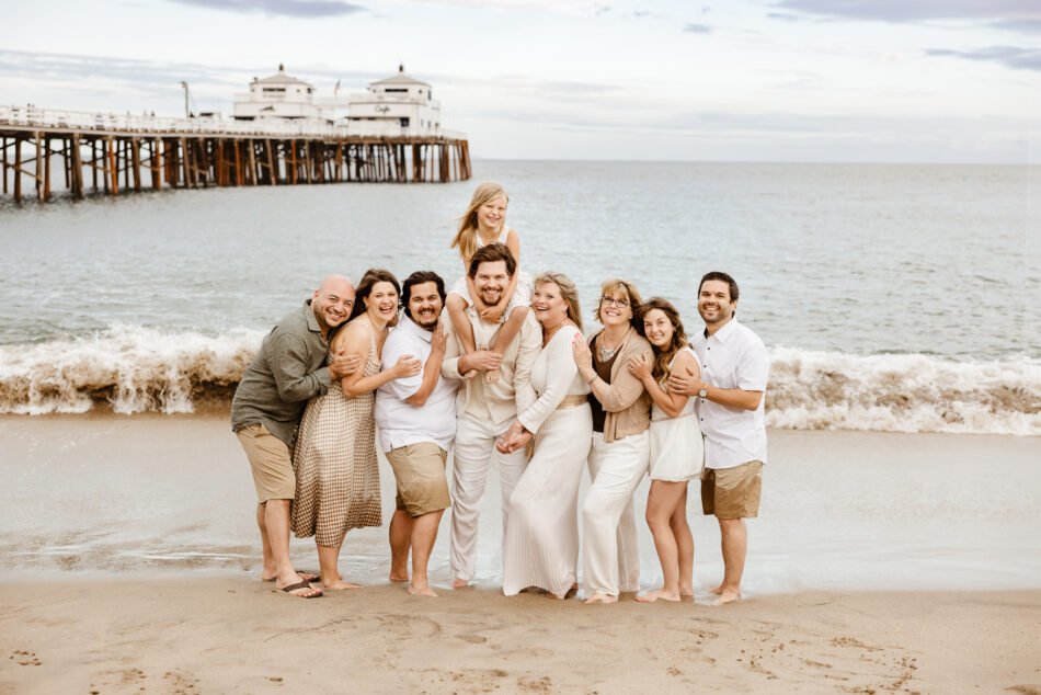 Extended family wearing white and neutral clothing posing together on the beach near the historic Malibu Pier.
