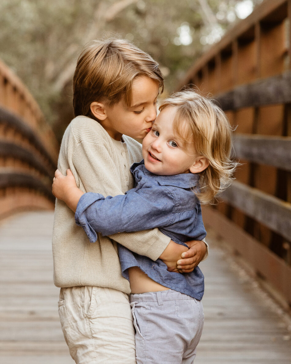 Older brother hugging and kissing his younger toddler brother on a wooden bridge during a Malibu family portrait session by Erica Montgomery Photography.