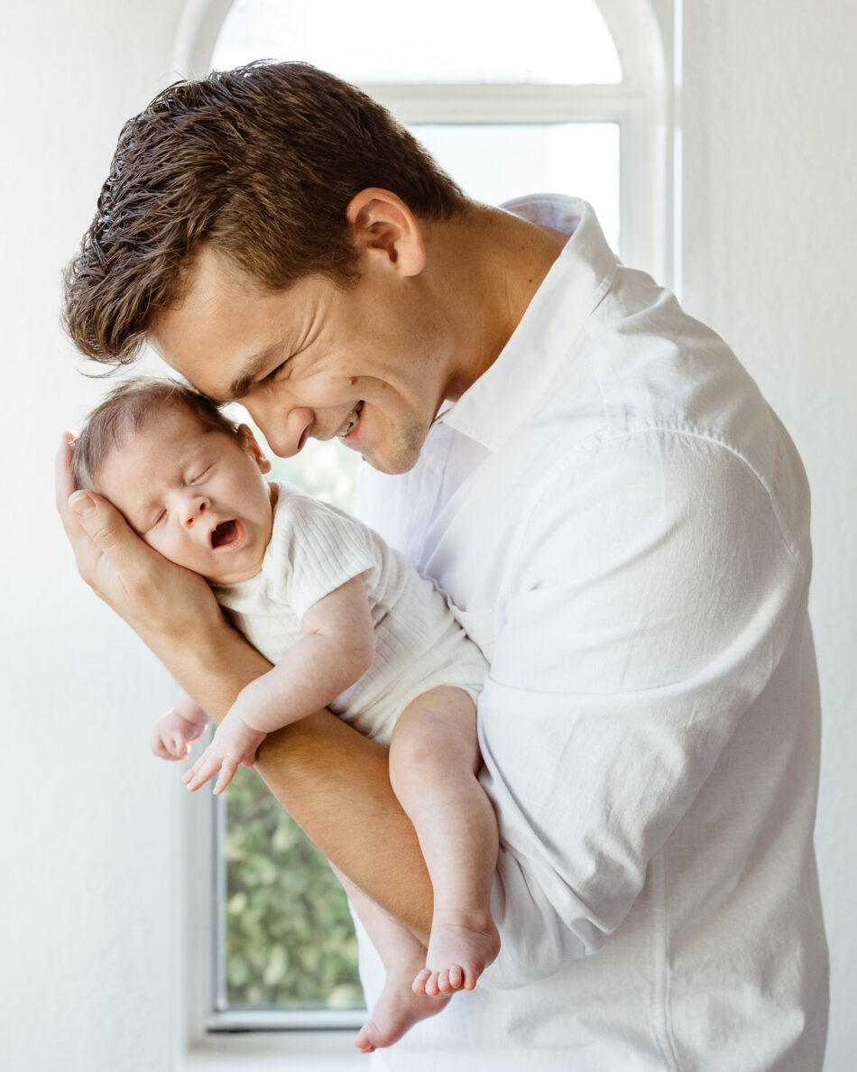Father cradling and smiling down at his yawning newborn baby during a Malibu home lifestyle photography session by Erica Montgomery Photography.