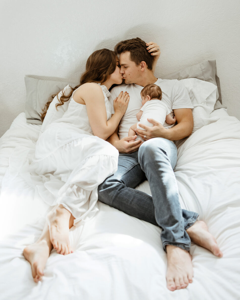 Parents cuddling and kissing on a white bed with their newborn baby during a relaxed Agoura Hills home photoshoot by Erica Montgomery Photography.