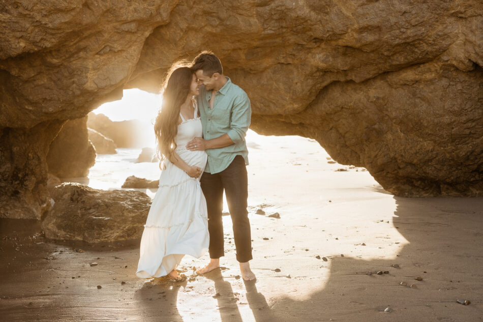 A wide-angle maternity photography session at sunset in Malibu, California. A pregnant woman in a white dress and her partner stand hand-in-hand, framed inside a large, dramatic coastal rock archway. Warm golden hour sunlight streams from behind them, casting long shadows on the sand and highlighting the rugged cliffs.