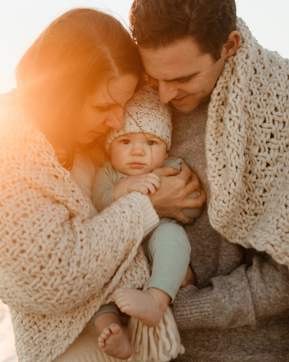 Parents snuggling and looking down at their newborn baby wrapped in cozy knit blankets during a Malibu family lifestyle photography session by Erica Montgomery Photography.