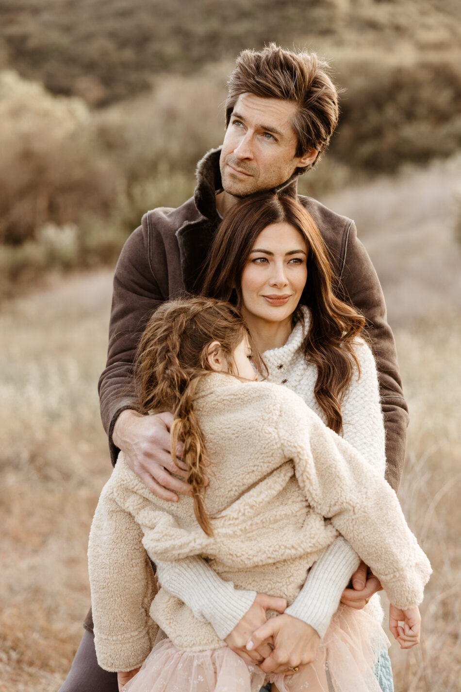Parents hugging their young daughter tightly from behind in an earthy canyon landscape during an Agoura Hills family photography session by Erica Montgomery Photography.