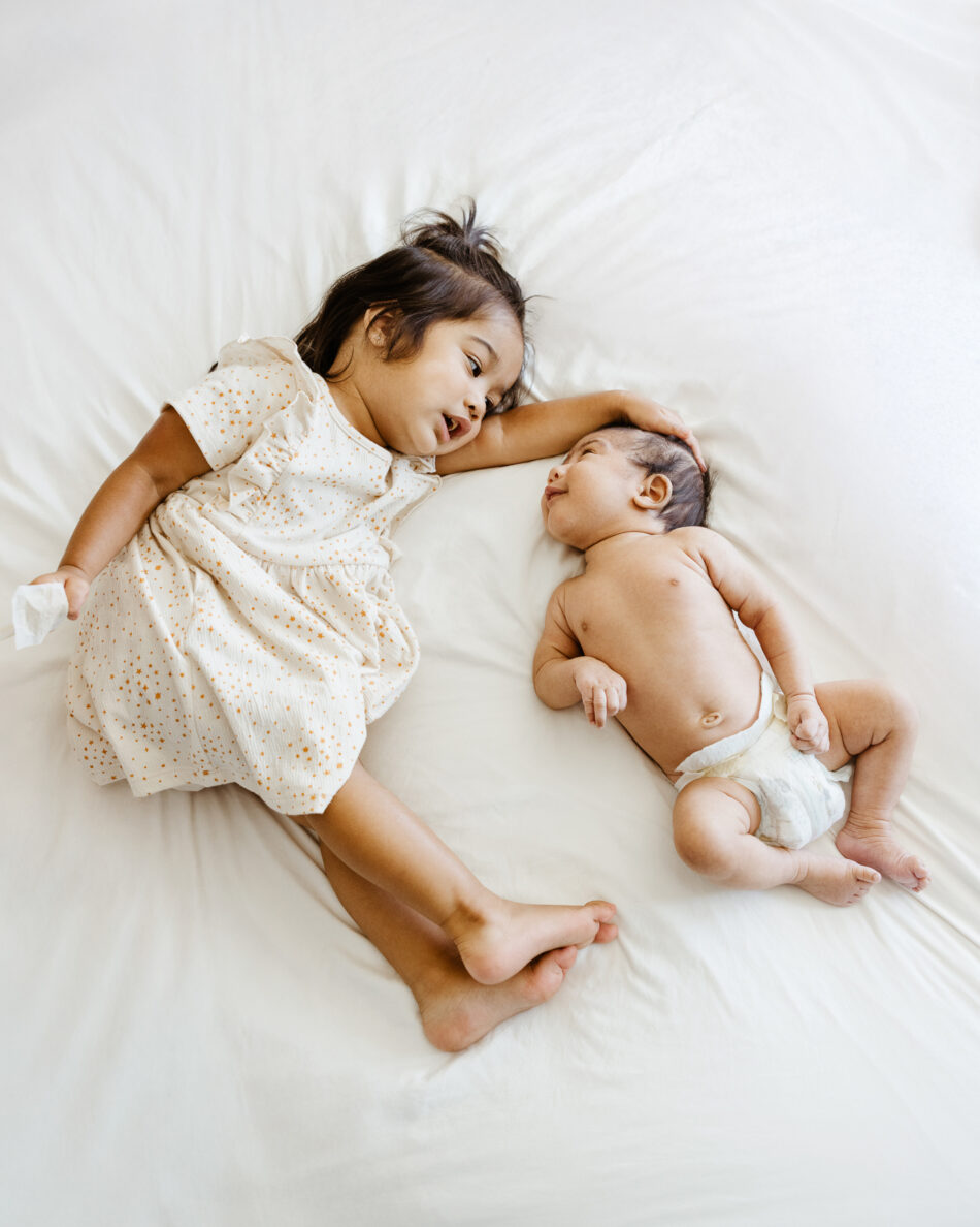 Big sister laying next to her newborn baby sibling on a white bed during a Malibu family lifestyle session by Erica Montgomery Photography.