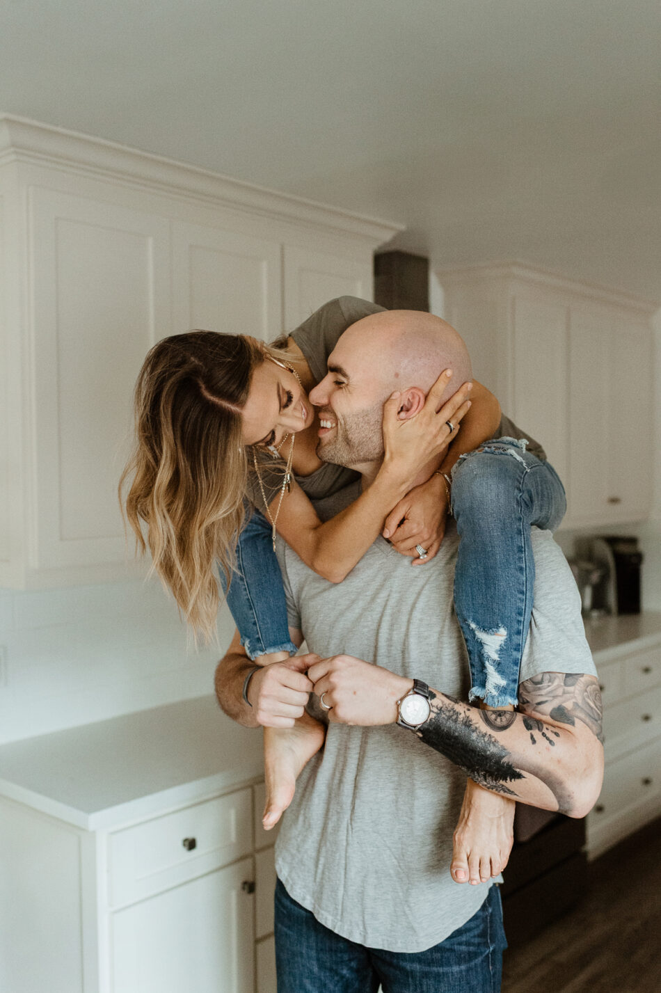 Playful in-home lifestyle couples photoshoot of a woman riding piggyback on her partner in a white kitchen by Erica Montgomery Photography.