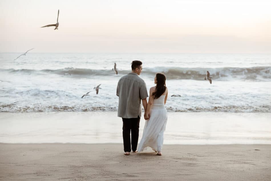 Engaged couple holding hands walking along the ocean shoreline with seagulls during a Malibu beach proposal session by Erica Montgomery Photography.