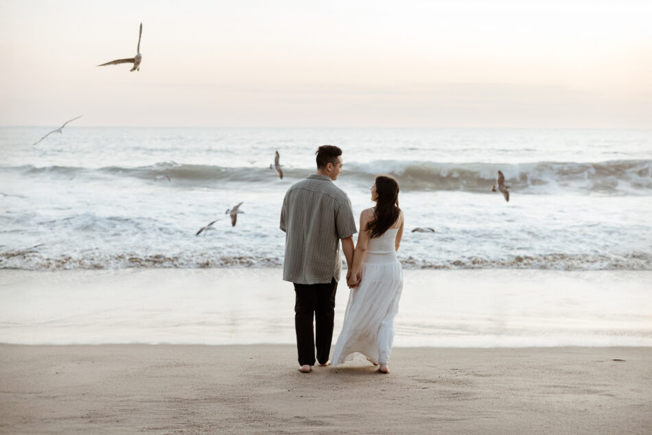 Engaged couple holding hands walking along the ocean shoreline with seagulls during a Malibu beach portrait session by Erica Montgomery Photography.