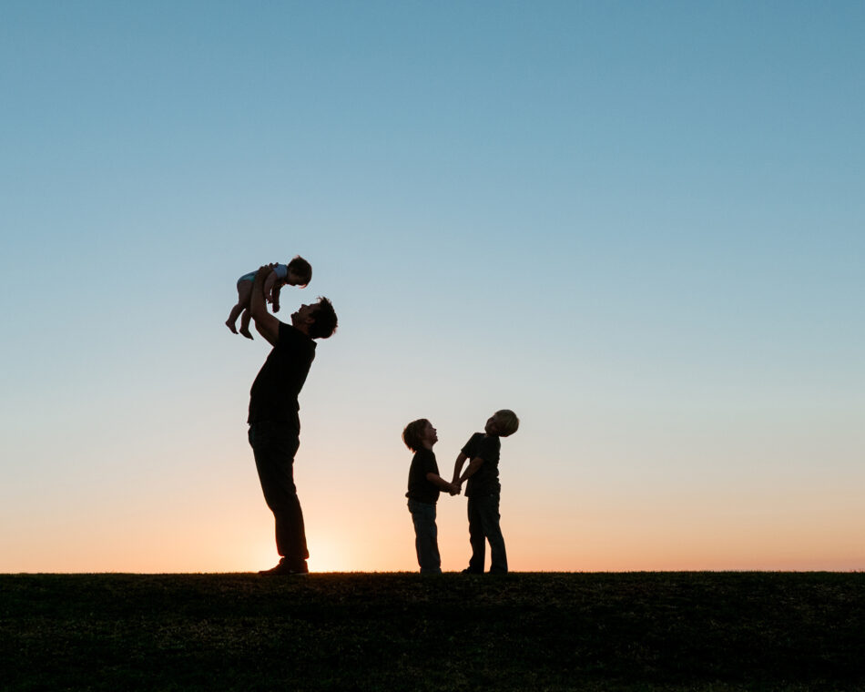 A dramatic sunset silhouette family photography session at Pepperdine University in Malibu, California. A father stands on a hill holding his toddler up in the air, while his two older children hold hands beside him against a clear, colorful twilight sky.