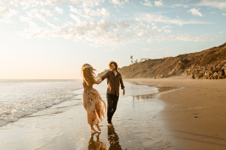 Man holding hands and spinning his fiancée along the shoreline during a cinematic Malibu beach engagement session by Erica Montgomery Photography.