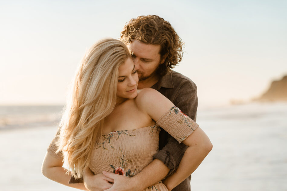Romantic beach engagement photos of a man hugging his blonde fiancée from behind in Malibu by Erica Montgomery Photography.