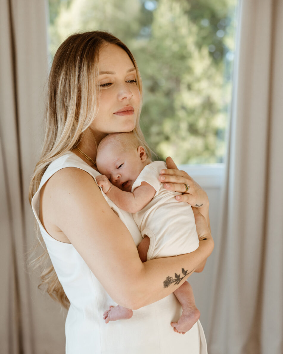 Close-up of a blonde mother tenderly holding her sleeping newborn baby by a window in Calabasas by Erica Montgomery Photography.