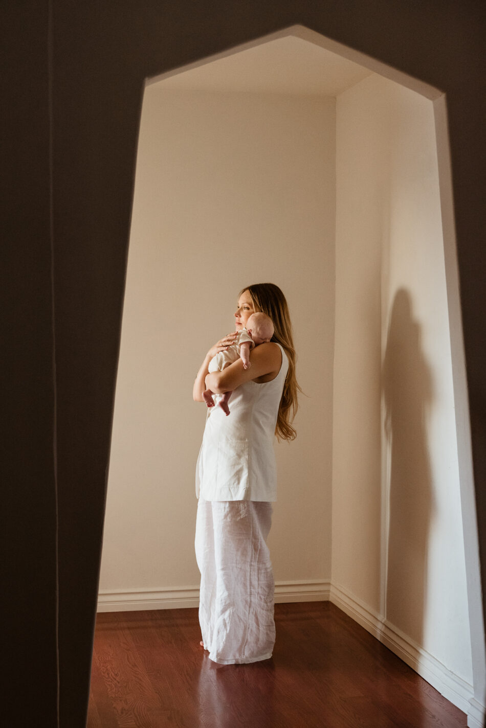 Artistic, framed portrait of a mother holding her newborn baby in a naturally lit architectural home hallway in Woodland Hills by Erica Montgomery Photography.