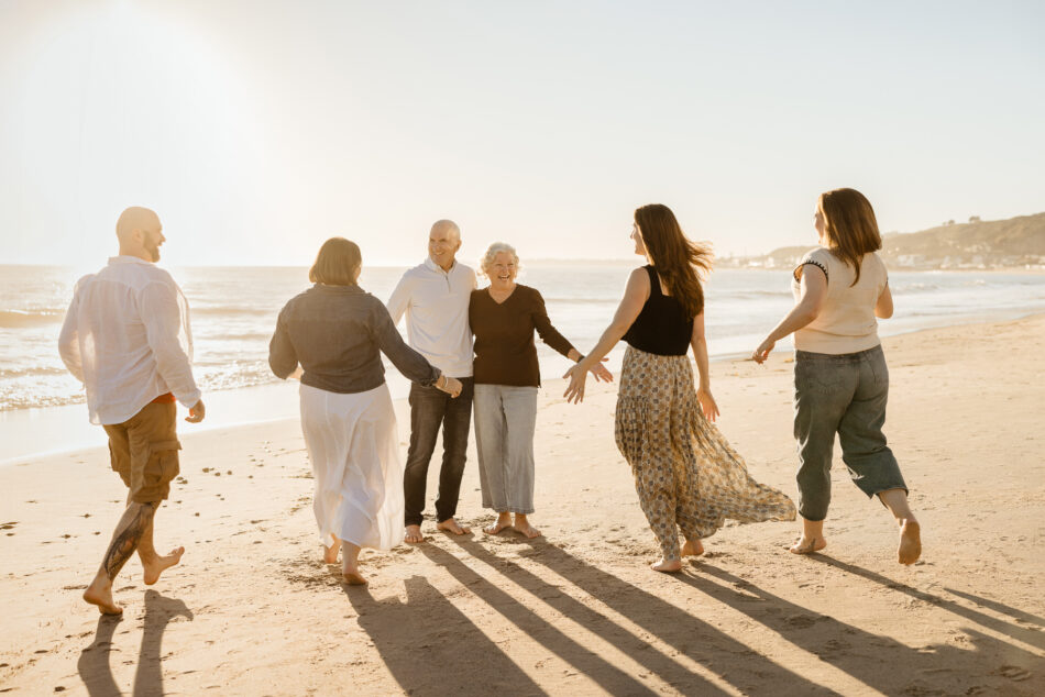 Candid editorial style photo of adult children running to their older parents on the sand with long shadows during a Malibu beach sunset.
