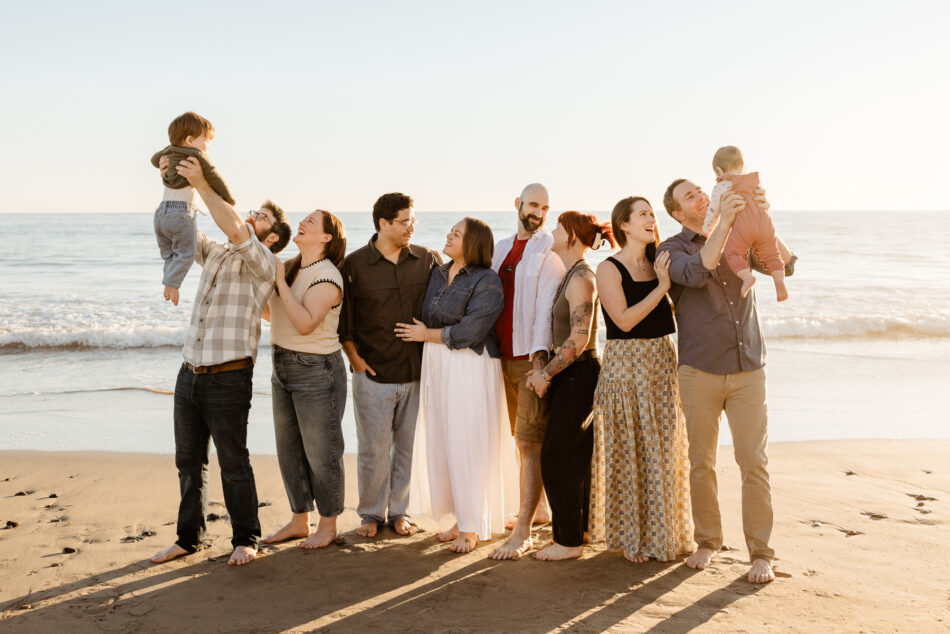 Large multi-generational family lining up on dry sand at golden hour for a Malibu vacation photography session and lifting babies in the air.
