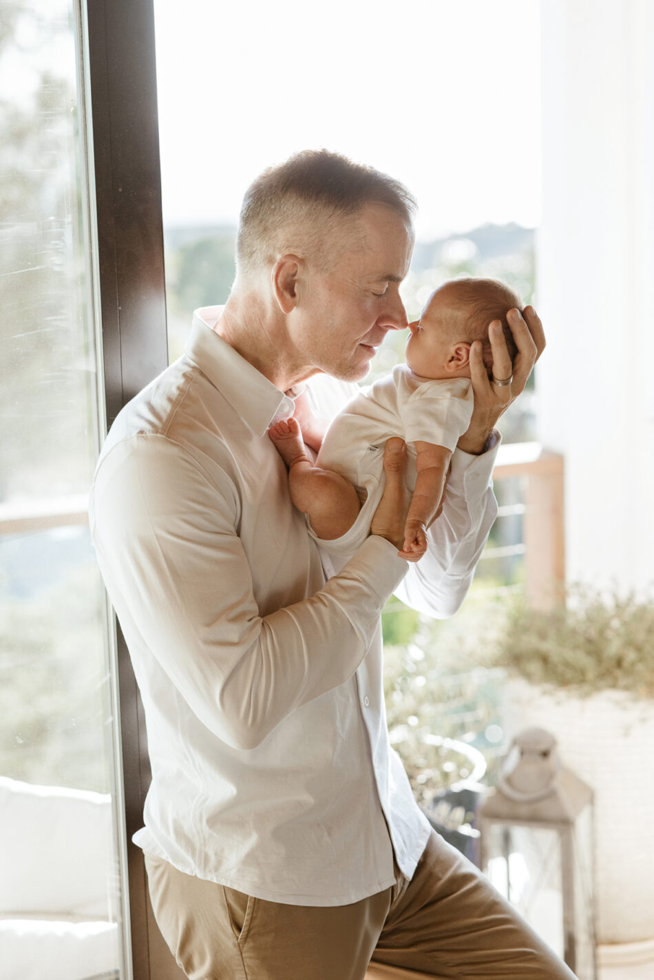 Father holding his newborn baby up nose-to-nose by a large bright window in Malibu by Erica Montgomery Photography.