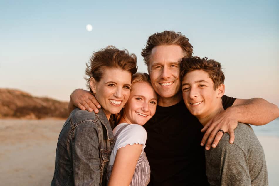 A warm, joyful family photography session on the beach in Malibu, California. A loving mother, father, daughter, and son stand closely wrapped in a tight embrace, smiling and laughing directly at the camera during golden hour.