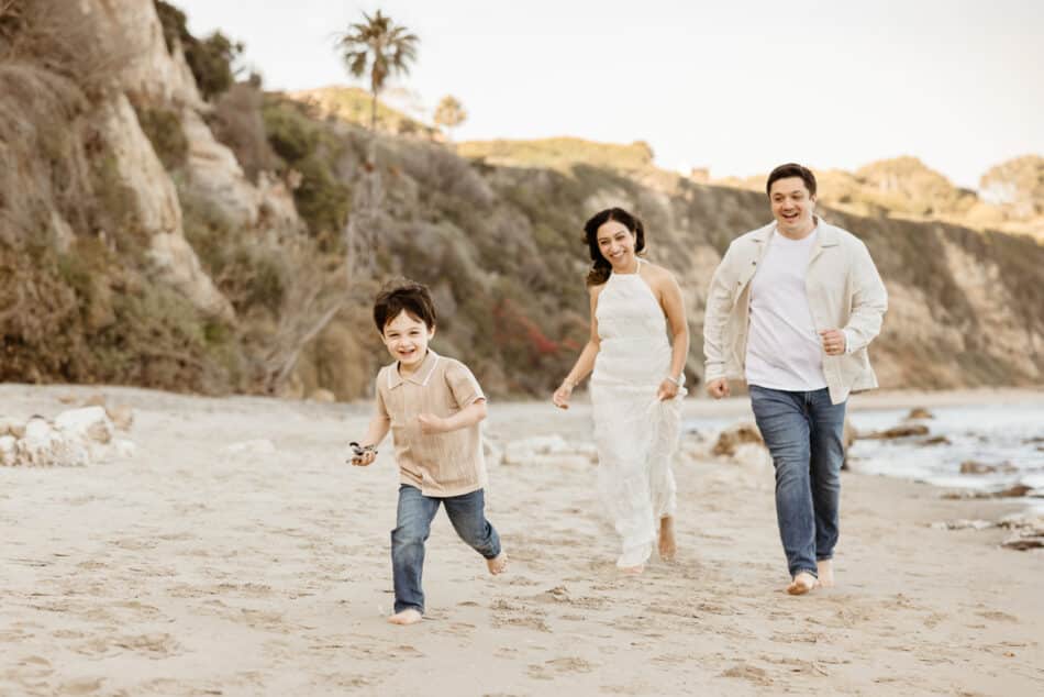 A candid family photography session at Point Dume in Malibu, California. A mother, father, and their young son laugh as they run together across the sandy beach during golden hour, with scenic ocean cliffs and a palm tree in the background.