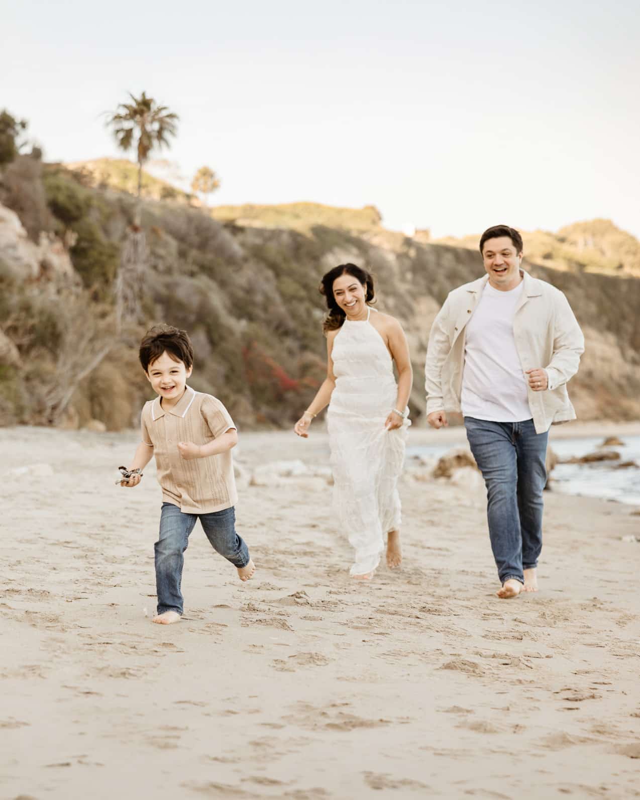 A candid family photography session at Point Dume in Malibu, California. A mother, father, and their young son laugh as they run together across the sandy beach during golden hour, with scenic ocean cliffs and a palm tree in the background.