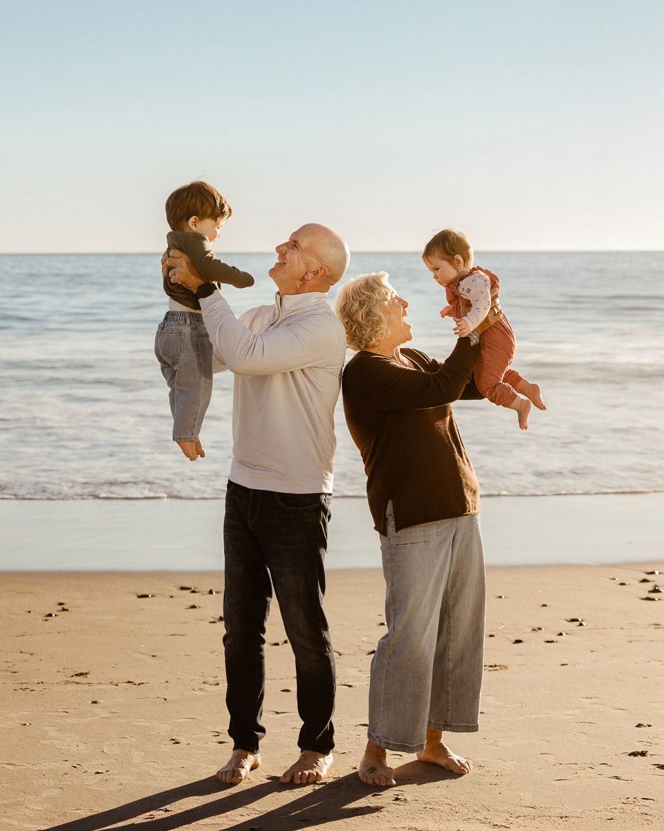 Grandparents holding up their baby grandchildren during an effortless beachside family reunion session at Malibu Colony Beach.